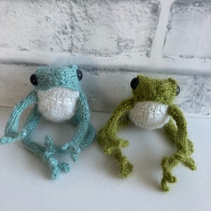 May include: Two small, handmade knitted frogs. One frog is light blue with a white belly, and the other is olive green with a white belly. Both frogs feature black plastic bead eyes and long, thin, articulated limbs with detailed feet.