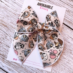 May include: A large, fabric hair bow featuring a repeating pattern of cartoon dog faces in various colors on a light beige background. The bow is on a white card with the text "ANORAHBLE bows". The bow is a handmade accessory.