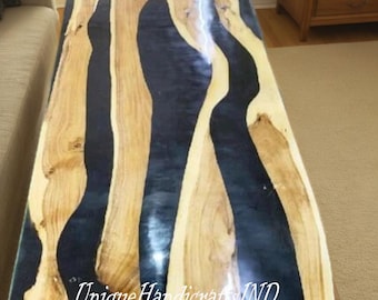 May include: A rectangular coffee table with a unique design. The tabletop features a combination of light brown wood and dark blue resin, creating a flowing pattern. The table is supported by black legs, and the text "Unique Handicrafts IND" is visible on the surface.