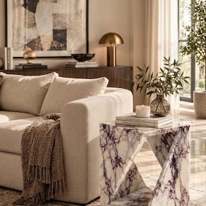 May include: A square side table crafted from white marble with prominent purple veining, featuring a geometric cutout design. The table is positioned next to a beige sofa draped with a brown textured throw blanket, with books and a glass vase on top.