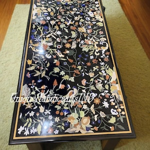 Op de afbeelding: Een zwarte salontafel met een inlegmotief van bloemen. De inleg is gemaakt van verschillende gekleurde stenen en heeft een verscheidenheid aan bloemen, bladeren en vogels. De tafel heeft een zwart houten frame en een gouden rand rond de rand van de inleg.