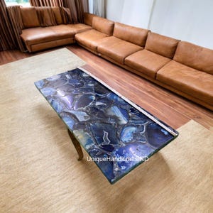 May include: A rectangular coffee table with a blue and purple agate stone top. The table has a unique pattern with a polished finish. The legs are a light brown color. The table is in a living room setting with a brown leather sofa and a beige rug.