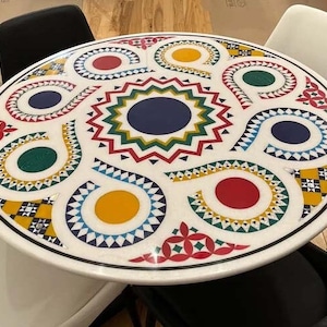 Puede incluir: Una mesa redonda blanca con un dise&ntilde;o geom&eacute;trico colorido. El dise&ntilde;o presenta colores rojo, amarillo, azul, verde y negro. La mesa est&aacute; hecha de m&aacute;rmol y tiene un acabado brillante.