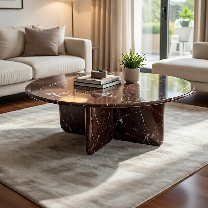 May include: A round, dark red marble coffee table with white veining. The table has a solid base and is placed on a light gray rug. A small potted plant and books are on the table.