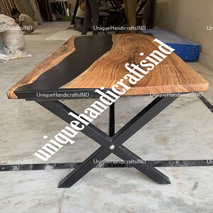 Op de afbeelding: Een rechthoekige houten tafel met een zwart epoxyhars rivierontwerp. De tafel heeft een natuurlijke houtnerf afwerking en wordt ondersteund door zwarte metalen kruispoten. De tafel is ongeveer 183 cm lang.