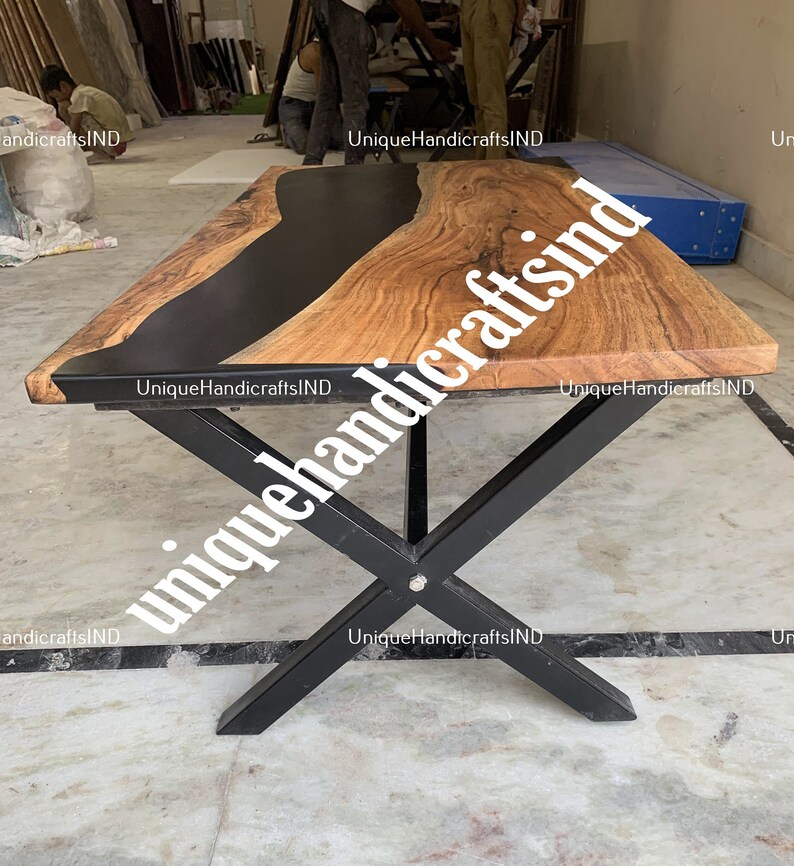 Op de afbeelding: Een rechthoekige houten tafel met een zwart epoxyhars rivierontwerp. De tafel heeft een natuurlijke houtnerf afwerking en wordt ondersteund door zwarte metalen kruispoten. De tafel is ongeveer 183 cm lang.