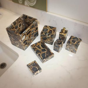 Puede incluir: Un juego de accesorios de baño con un patrón único. El juego incluye un porta cepillos de dientes rectangular, un jabonera, una caja cuadrada pequeña, un dispensador de jabón, un pequeño recipiente cuadrado y otros dos recipientes. Los accesorios tienen un patrón marrón, negro y blanco.