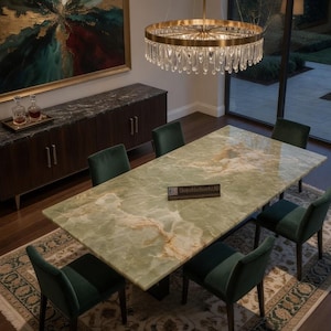 Op de afbeelding: Een eethoek met een rechthoekige groene marmeren tafel en zes groene fluwelen stoelen. Een donkere houten kast en een groot abstract schilderij zijn zichtbaar. Boven de tafel hangt een gouden kroonluchter.