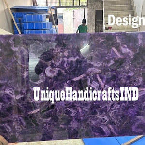 May include: A close-up of a purple amethyst slab with a swirling pattern. The slab is being held by a person's hand. The text "UniqueHandicraftsIND" is visible in the lower center of the image. The text "Design 1" is visible in the upper right corner of the image.