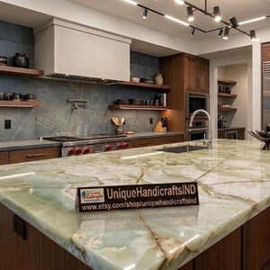 May include: A modern kitchen with a large island featuring a green and white marble countertop. The kitchen has dark wood cabinetry, stainless steel appliances, and a range hood. Shelves display decorative items.