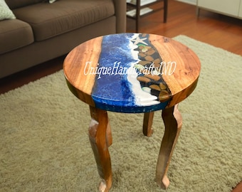May include: Round wooden side table with a blue and white epoxy resin top featuring a beach scene with seashells and stones. The table has three legs.