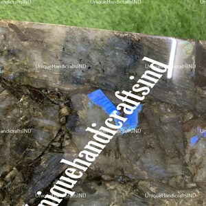 May include: Close-up of a dark blue and grey labradorite stone with iridescent blue and green hues. The stone has a rough, natural texture. The image is watermarked with the text "Unique HandicraftsIND".