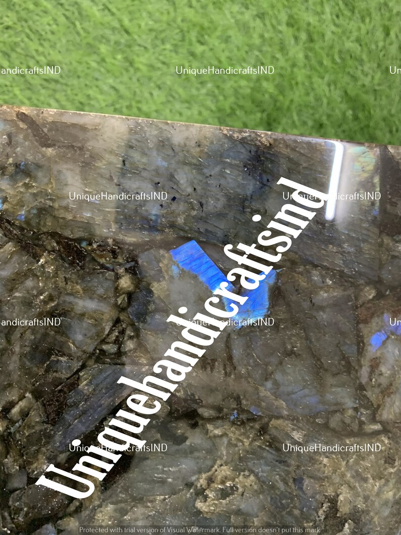 May include: Close-up of a dark blue and grey labradorite stone with iridescent blue and green hues. The stone has a rough, natural texture. The image is watermarked with the text "Unique HandicraftsIND".
