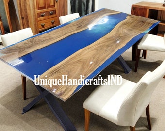 Puede incluir: Una mesa de comedor de madera con una incrustación de resina epoxi azul. La mesa tiene una base en forma de X de metal negro y está rodeada de sillas blancas. La mesa está etiquetada "UniqueHandicraftsIND".