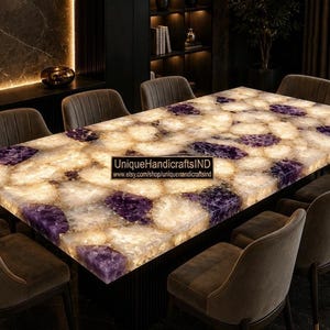 May include: A rectangular dining table featuring a backlit surface made of inlaid purple amethyst and translucent white stone segments. The table has a smooth, polished top and is surrounded by eight upholstered chairs. Text reads UniqueHandicraftsIND.
