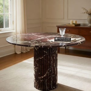 Puede incluir: Una mesa redonda de mármol con una superficie con vetas rojas oscuras y blancas y una base cilíndrica. La mesa tiene unos 100 cm de diámetro. Hay un vaso y un pequeño letrero sobre la mesa.