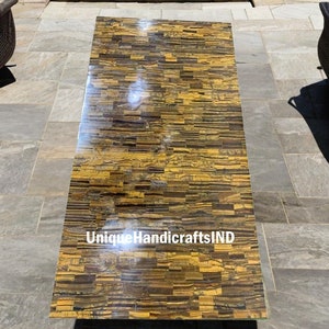 May include: A rectangular table top made of polished tiger's eye stone. The table top is made of many small pieces of tiger's eye stone that are arranged in a pattern. The table top is a rich golden brown color with black and gold streaks. The text "UniqueHandicraftsIND" is visible on the table top.