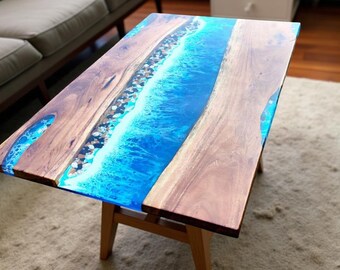 May include: A rectangular coffee table with a wooden frame and top. The table's surface features a central blue resin design resembling ocean waves, flanked by sections of natural wood. The table is supported by four wooden legs.