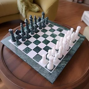 Könnte beinhalten: Ein Schachspiel mit einem grün-weiß karierten Brett und Schachfiguren. Die Schachfiguren sind weiß und dunkelgrün. Das Brett steht auf einem runden, dunkelbraunen Holztisch.