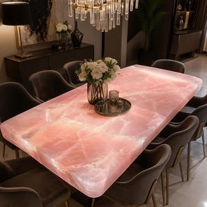 May include: A large, rectangular pink dining table with a glowing, translucent surface. The table is surrounded by eight gray velvet chairs with gold legs. A chandelier hangs above, and a vase of white flowers sits on the table.