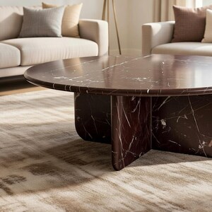May include: A round, dark brown marble coffee table with white veining. The table has a thick, rounded top and a pedestal base with four curved supports. The table is in a living room setting with neutral-toned furniture and a patterned rug.