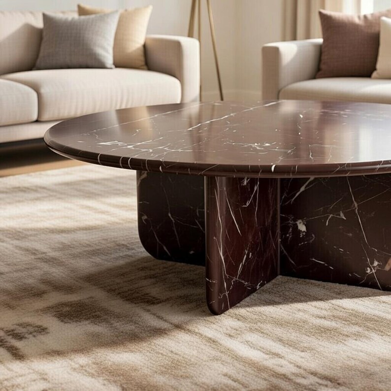 May include: A round, dark brown marble coffee table with white veining. The table has a thick, rounded top and a pedestal base with four curved supports. The table is in a living room setting with neutral-toned furniture and a patterned rug.