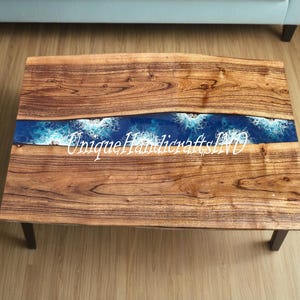 Op de afbeelding: Een rechthoekige houten salontafel met een donkerblauw hars rivierontwerp. Het rivierontwerp heeft witte golfachtige patronen. De tafel heeft donkerbruine poten en de tekst "Unique Handicrafts IND" is zichtbaar.