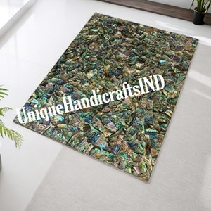 Puede incluir: Una alfombra iridiscente verde, azul y dorada hecha de nácar. La alfombra está hecha de pequeñas piezas irregulares de concha que están dispuestas en un patrón aleatorio. La alfombra tiene un aspecto único y elegante. El texto "Unique Handicrafts IND" es visible en la imagen.