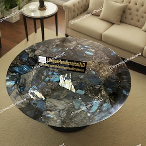 May include: A round, dark blue and black coffee table with a unique, mosaic-like top made of labradorite stone. The table is sitting on a beige rug in a living room setting.