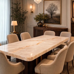 Op de afbeelding: Een rechthoekige eettafel met een gepolijst lichtbeige stenen blad met een opvallende amberkleurige ader, ondersteund door een donkerbruine gecanneleerde voet, omringd door acht gestoffeerde beige stoelen met decoratieve nagelafwerking.