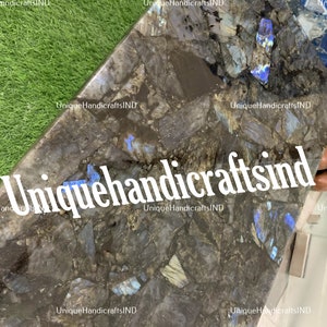 May include: A close-up of a dark grey and blue table top made of labradorite stone. The stone has a unique iridescent quality, with flashes of blue, green, and gold. The table top is cut in a geometric shape, with sharp angles and edges. The text "Uniquehandicraftsind" is visible in the image.