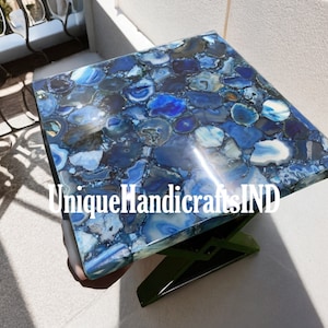 Könnte beinhalten: Blaue Achat-Tischplatte mit einem einzigartigen Muster aus blauen und weißen Achat-Scheiben. Die Tischplatte wird von einem schwarzen X-förmigen Gestell getragen. Der Text "UniqueHandicraftsIND" ist auf der Tischplatte sichtbar.