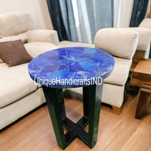 Op de afbeelding: Een ronde bijzettafel met een blauw lapis lazuli stenen blad en een zwarte metalen basis. De tafel staat in een woonkamer met een witte bank en een houten vloer.