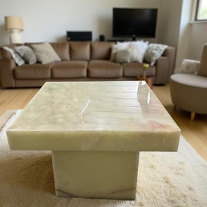 May include: A square, light green onyx coffee table with a rectangular base. The table has a polished surface and sits on a beige rug. The table is in a living room setting.