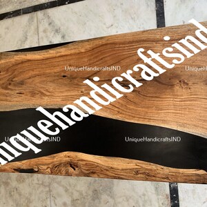Op de afbeelding: Een rechthoekige houten tafel met een donkere, glanzende zwarte harsrivier in het midden. Het hout heeft een natuurlijk nerfpatroon met warme bruine tinten. De tekst "uniquehandcraftsind" is over de afbeelding geplaatst.