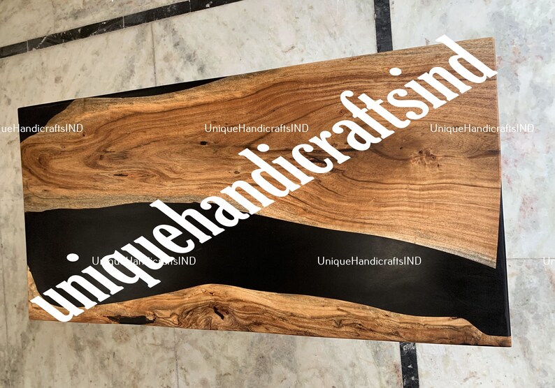 Op de afbeelding: Een rechthoekige houten tafel met een donkere, glanzende zwarte harsrivier in het midden. Het hout heeft een natuurlijk nerfpatroon met warme bruine tinten. De tekst "uniquehandcraftsind" is over de afbeelding geplaatst.