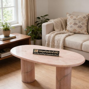 May include: An oval coffee table crafted from polished pink marble with two cylindrical pedestal bases. A small rectangular black sign sits on the tabletop featuring the text UniqueHandicraftsIND and www.etsy.com/shop/uniquehandicraftsind.