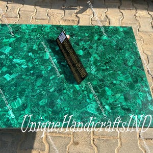 May include: A green malachite tabletop with a unique pattern of swirls and veins. The tabletop is made of natural stone and has a polished finish. The text "Unique Handicrafts IND" is visible in the bottom right corner of the image.
