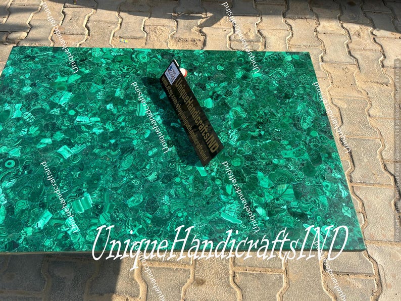 May include: A green malachite tabletop with a unique pattern of swirls and veins. The tabletop is made of natural stone and has a polished finish. The text "Unique Handicrafts IND" is visible in the bottom right corner of the image.