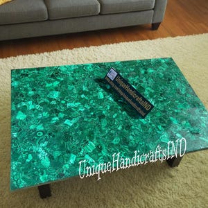 May include: A green malachite coffee table with a unique, natural pattern. The table is made of polished stone and has a rectangular shape. The table is sitting on a white rug. The text "Unique Handicrafts IND" is visible on the table.