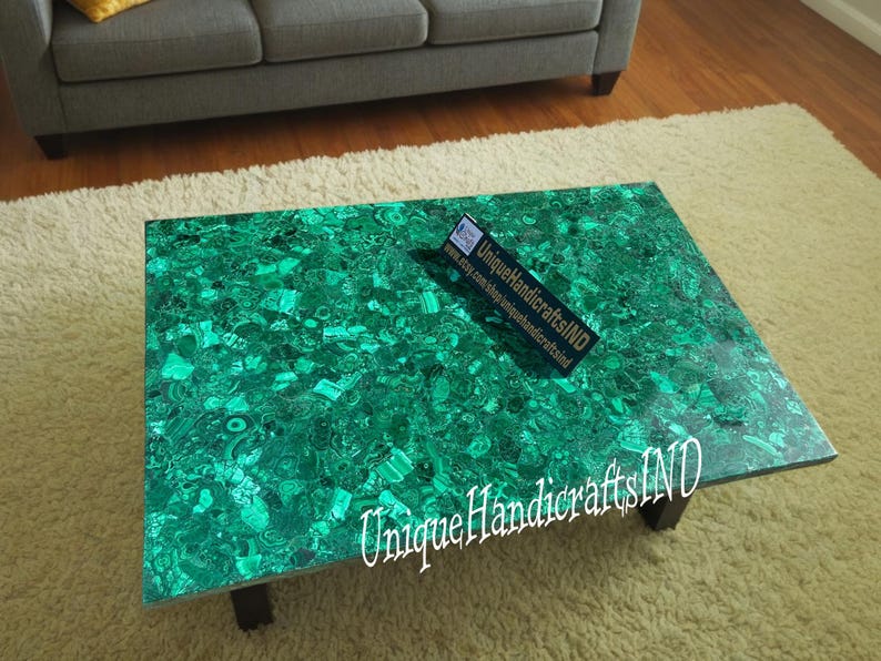 May include: A green malachite coffee table with a unique, natural pattern. The table is made of polished stone and has a rectangular shape. The table is sitting on a white rug. The text "Unique Handicrafts IND" is visible on the table.