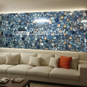 May include: A white sofa with orange and white pillows in front of a wall with a large blue agate stone wall art piece. The art piece has the text "Unique Handicrafts IND" on it.