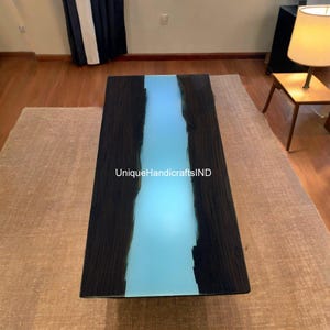 May include: A dark wood coffee table with a light blue epoxy river running down the center. The table is lit from underneath, making the epoxy glow. The text "UniqueHandicraftsIND" is visible on the table.