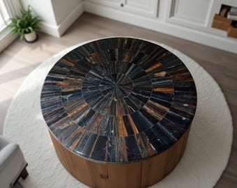 Op de afbeelding: Een ronde salontafel met een mozaïekblad met een radiaal patroon van blauwe, bruine en gouden stenen. De basis is gemaakt van lichtbruin hout. De tafel staat op een wit, rond tapijt.