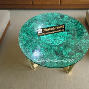 May include: A round green malachite table top with gold metal legs. The table top has a unique pattern of green and black swirls. The table is 30 inches in diameter. The table is on a beige carpet. The text "UniqueHandicraftsIND" is printed on the table top.