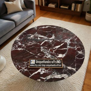 May include: A round coffee table with a dark red marble top featuring white veining. The table is set on a white shag rug. The table has the text "UniqueHandicraftsIND" and a website address.