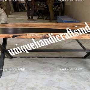 Op de afbeelding: Een rechthoekige houten tafel met een zwarte epoxyharsinlay en zwarte metalen poten. Het tafelblad is lichtbruin met donkere houtnerf. De poten zijn gekruist en schuin. De tekst "UniqueHandicraftsIND" is zichtbaar.
