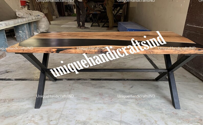 Op de afbeelding: Een rechthoekige houten tafel met een zwarte epoxyharsinlay en zwarte metalen poten. Het tafelblad is lichtbruin met donkere houtnerf. De poten zijn gekruist en schuin. De tekst "UniqueHandicraftsIND" is zichtbaar.