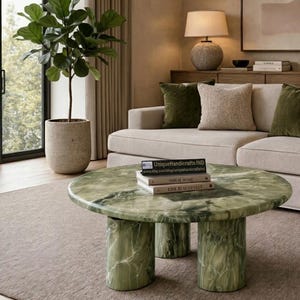 May include: A round green marble coffee table with a polished surface and three cylindrical legs. Two stacked books rest on the table, with the top book displaying the text "THIS IS HOME" and "LIVE BEAUTIFULLY" and a small black sign with white text.