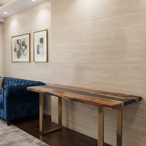 May include: A rectangular console table featuring a natural live-edge wood slab surface with a central black resin inlay. The table is supported by two brushed gold metal U-shaped legs, providing a stable base for the elongated wooden tabletop.